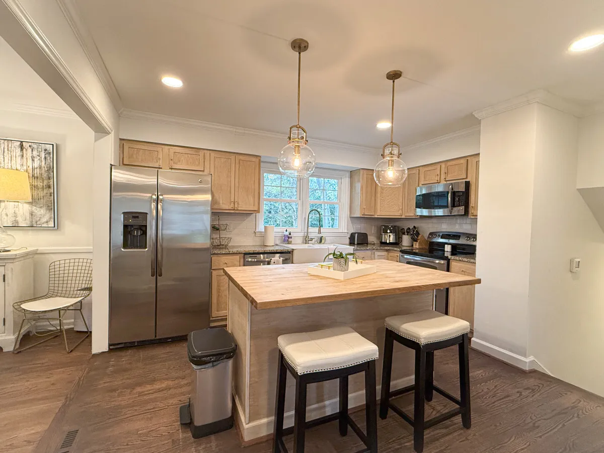 A modern kitchen illuminated by pendant lights, featuring an island countertop, stainless steel appliances, and a window letting in natural light. The scene feels inviting for meal prep or coffee breaks after a long workday.