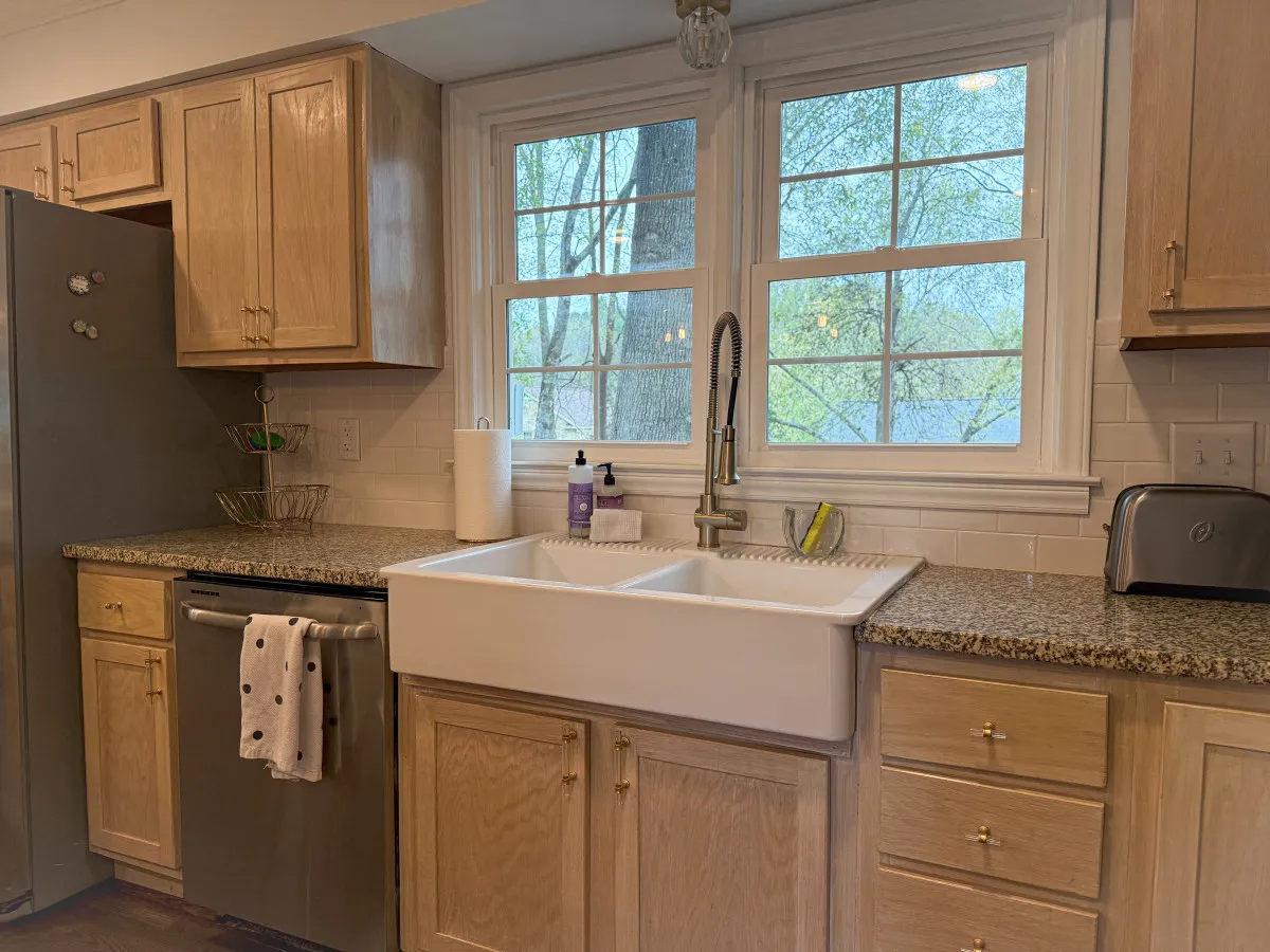 A bright kitchen featuring a large farmhouse sink under a window with treetop views, granite countertops, and stainless steel appliances. The space is airy and inviting, perfect for meal preparation and casual dining.