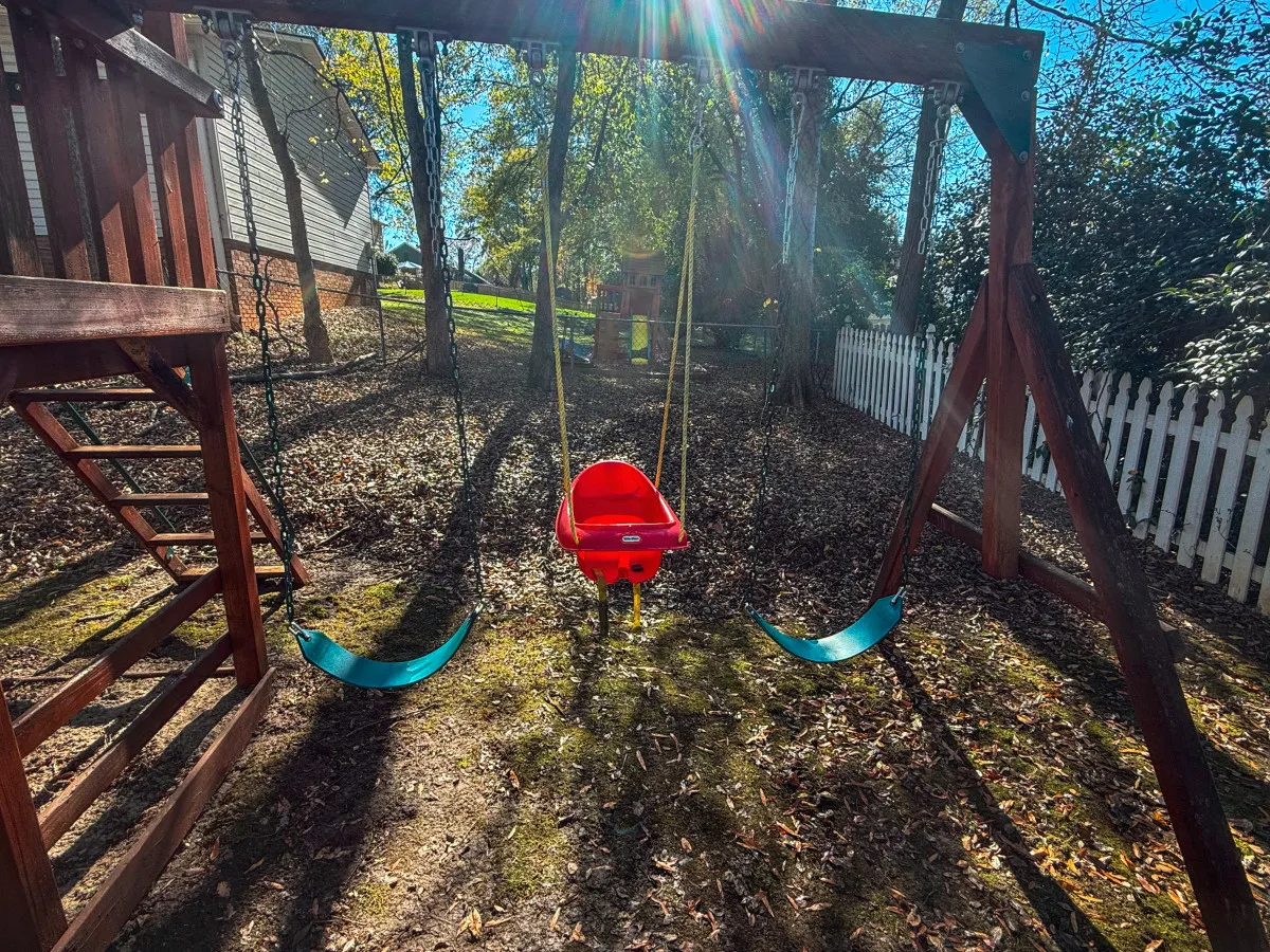 A cheerful outdoor setting with a wooden playset, red and green swings, and a white picket fence amidst autumn leaves. The scene radiates happiness and is ideal for family gatherings, playdates, and creating memories in a safe, fenced yard.