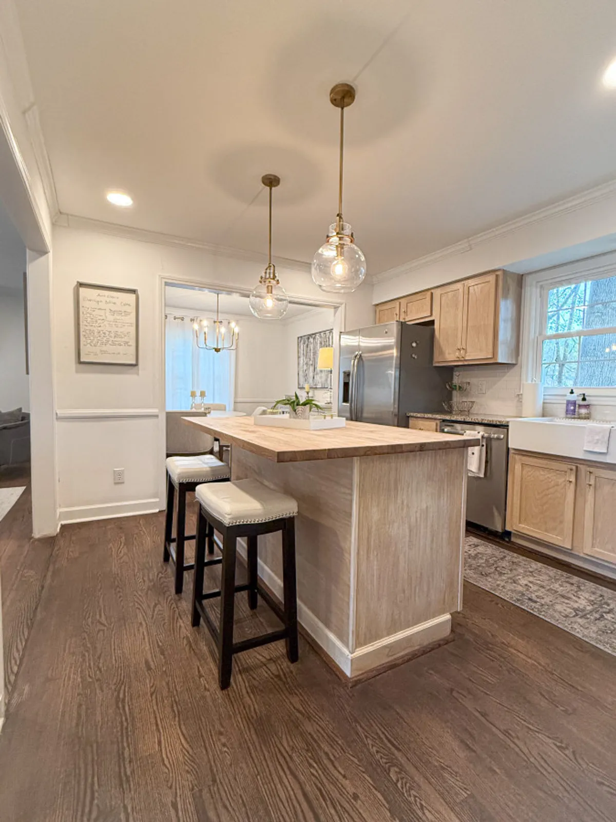 A spacious kitchen with a large island, pendant lighting, stools for seating, and abundant natural light streaming in from a window. Stainless steel appliances and welcoming decor set the tone for gatherings, cooking, and casual dining. The scene is ideal for both family meals and entertaining guests.