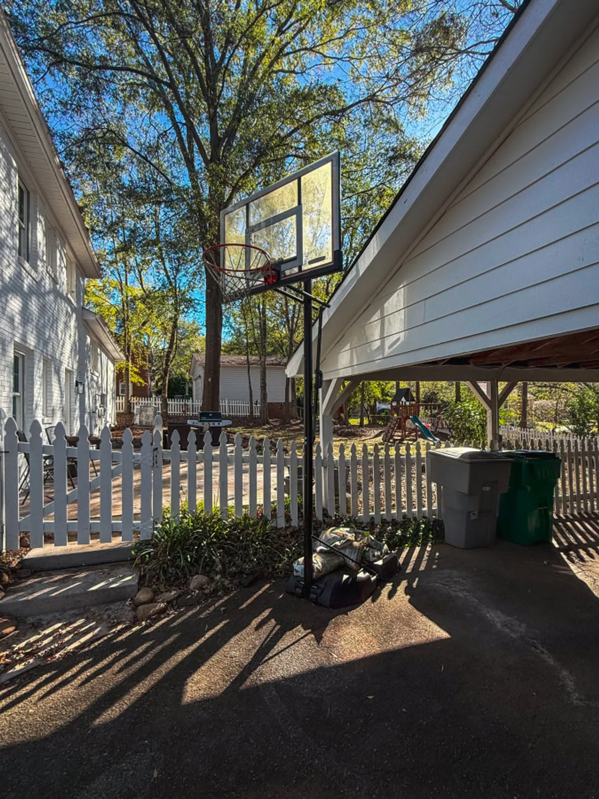 This photo captures a backyard scene with a basketball hoop, a white fence, trees, and playground equipment. The area is well-lit and inviting, suggesting possibilities for sports, play, and outdoor socializing in a safe, enclosed environment.