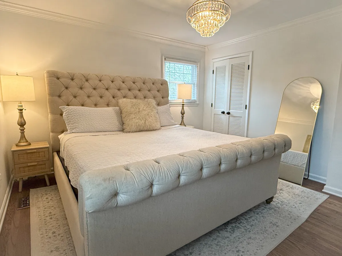 A luxurious bedroom featuring a plush bed with elegant bedding, a sparkling chandelier overhead, and a mirrored dresser. Soft natural light filters into the serene space, accentuating the carefully curated decor and creating a restful atmosphere.