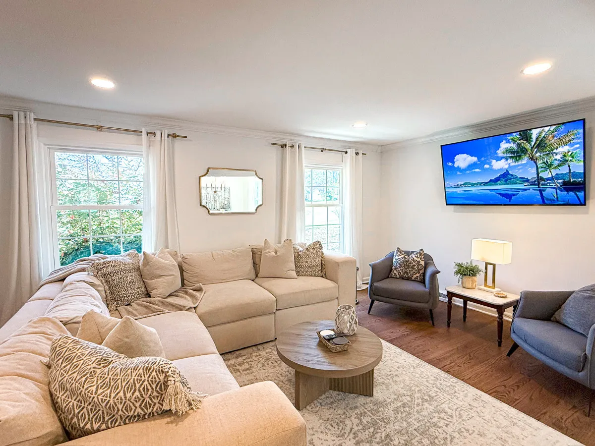 The image showcases a spacious and inviting living room with a large sectional sofa perfect for gatherings, stylish artwork on the wall, and a TV displaying a vibrant tropical scene. The setup encourages relaxation, group conversations, and movie nights, all within a cozy, well-lit space that's ideal for families or groups of friends.