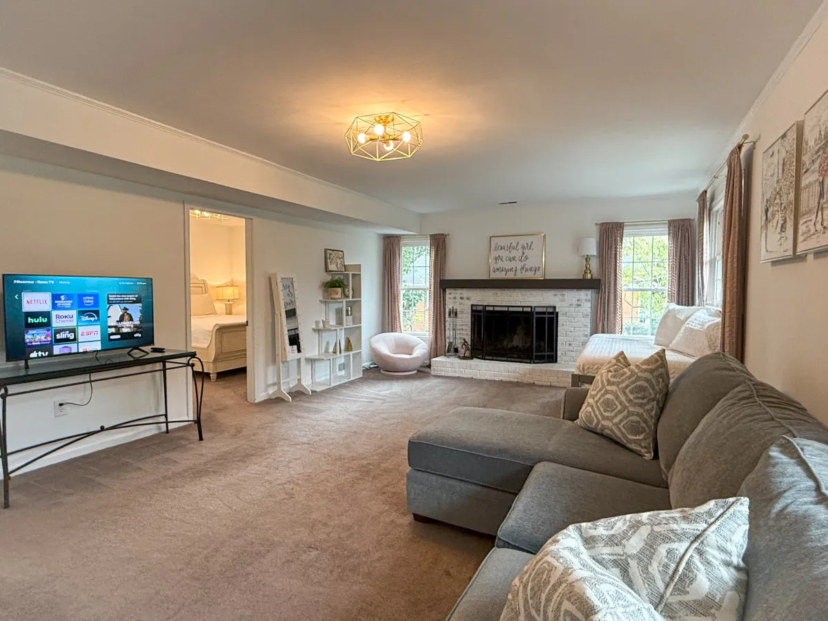 A cozy, professionally arranged living space featuring a sofa, fireplace, television, and artwork. Large windows allow natural light, highlighting the room's warmth and cleanliness. The scene is spacious yet intimate, reflecting a high standard of care.