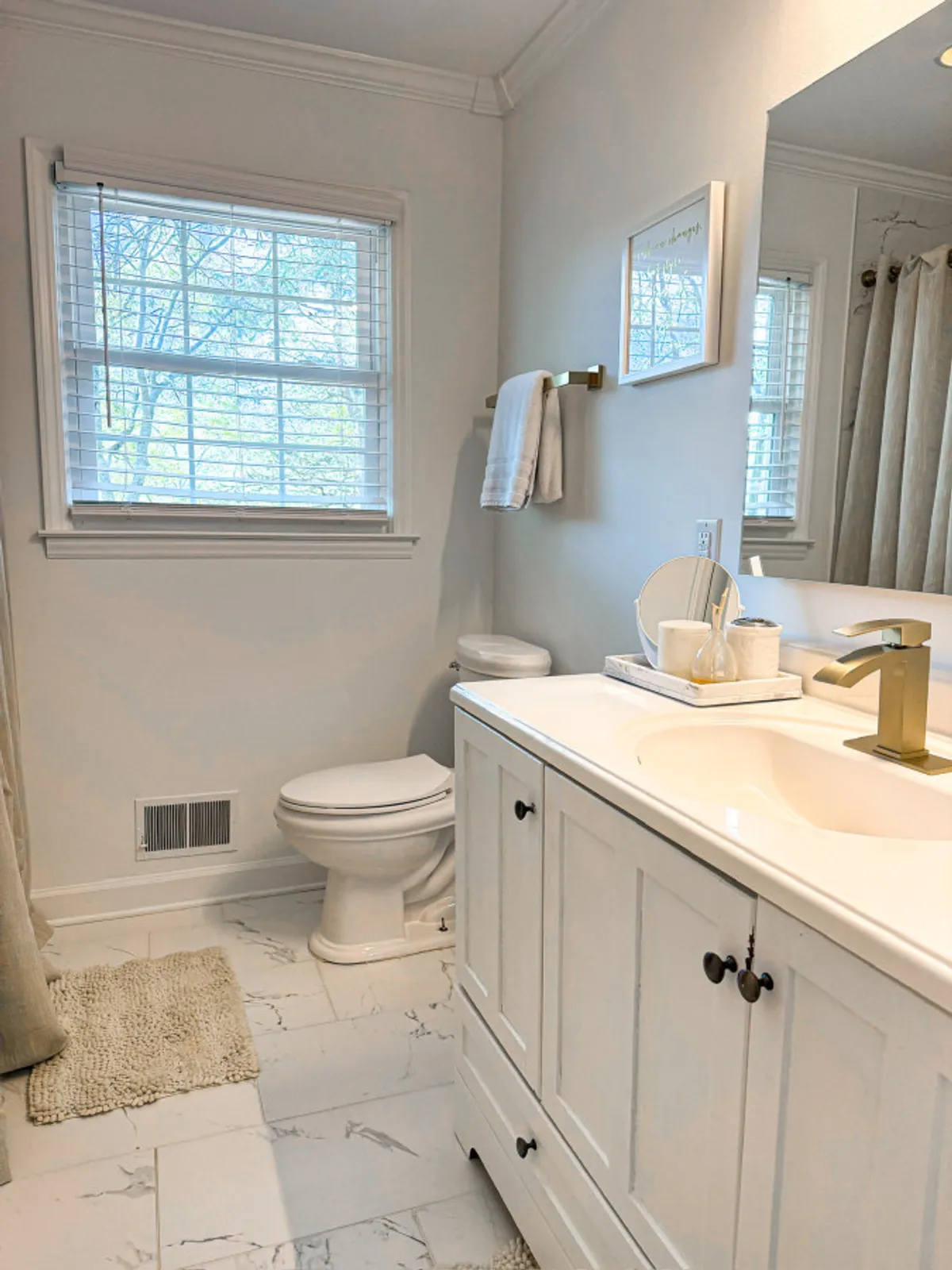 A modern bathroom with a well-lit vanity, mirror, towel rack, and a window with blinds. The surfaces are clean and inviting, offering the perfect setup for daily routines and self-care.