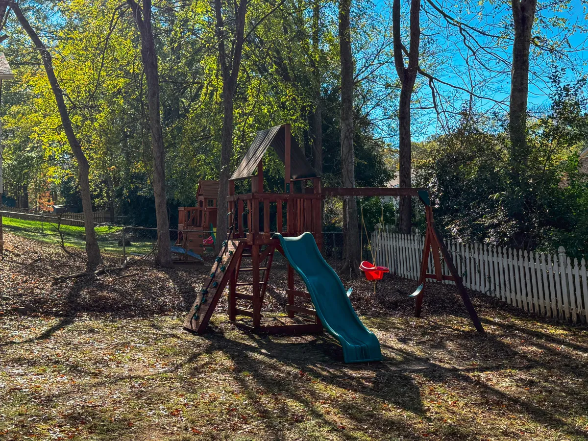 A playful backyard scene with a sturdy children's playset, including a swing and slide, set against a backdrop of mature trees and a secure fence. The shaded area and safe play surfaces make it an ideal spot for kids to enjoy carefree afternoons while adults relax nearby.