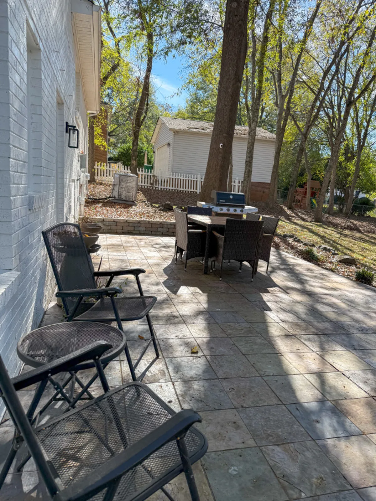 A casual and welcoming patio scene featuring a paved area with a black folding outdoor dining table, surrounded by green trees and ample space for socializing or relaxing. The setup is versatile, suitable for both alfresco meals and relaxed afternoons under the sun.