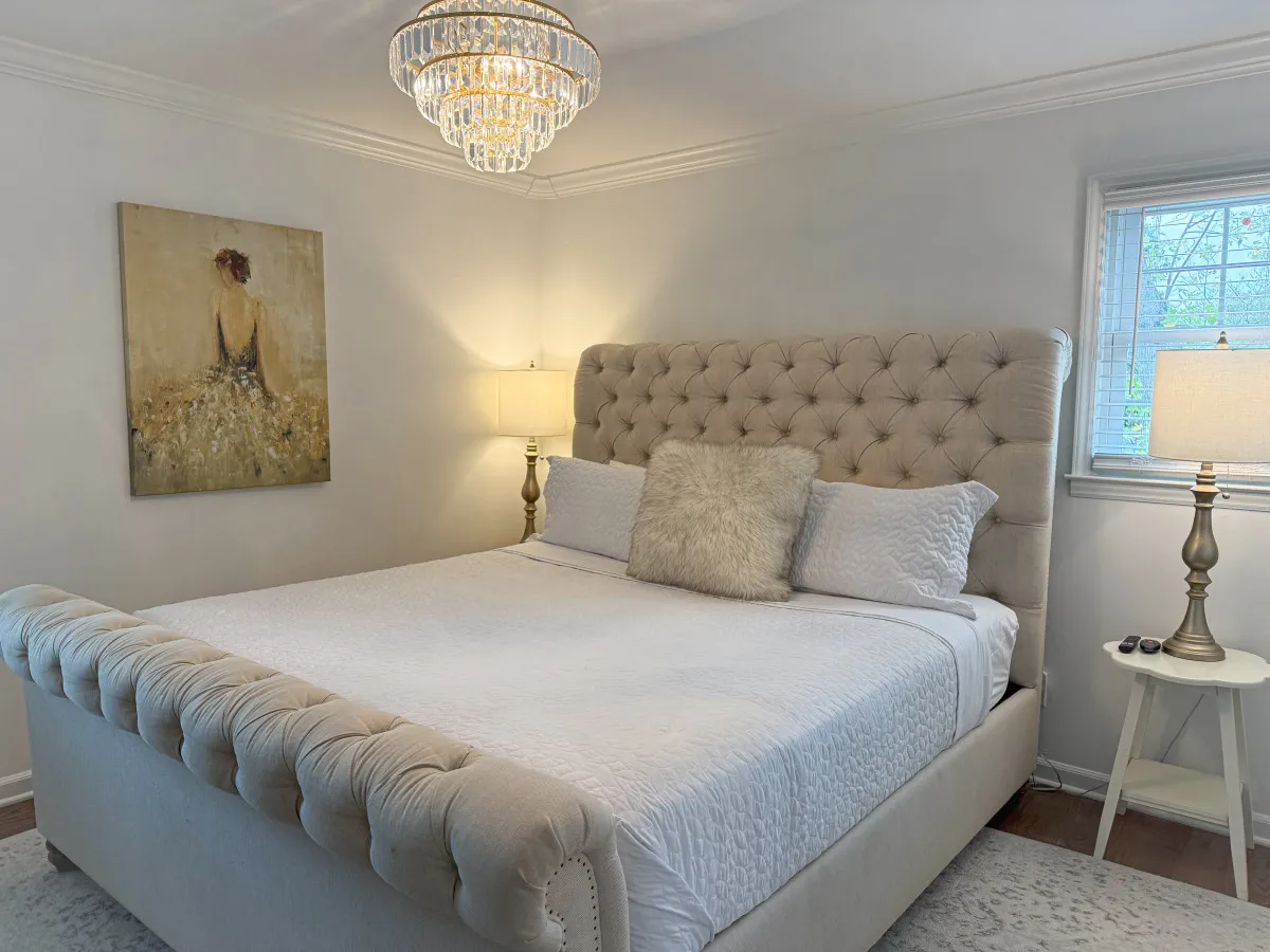 A peaceful bedroom highlighted by a chandelier, comfortable bed, tasteful artwork, and a nightstand with a lamp. The room radiates calmness and comfort, ideal for both restful sleep and quiet reading.