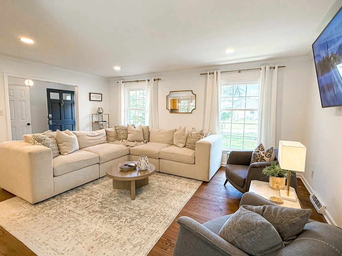 A cozy living room featuring a large sectional sofa, a round coffee table, a wall-mounted TV, and beautiful natural light pouring in through multiple windows. This inviting setup is ideal for movie nights, socializing, or relaxing as a family.