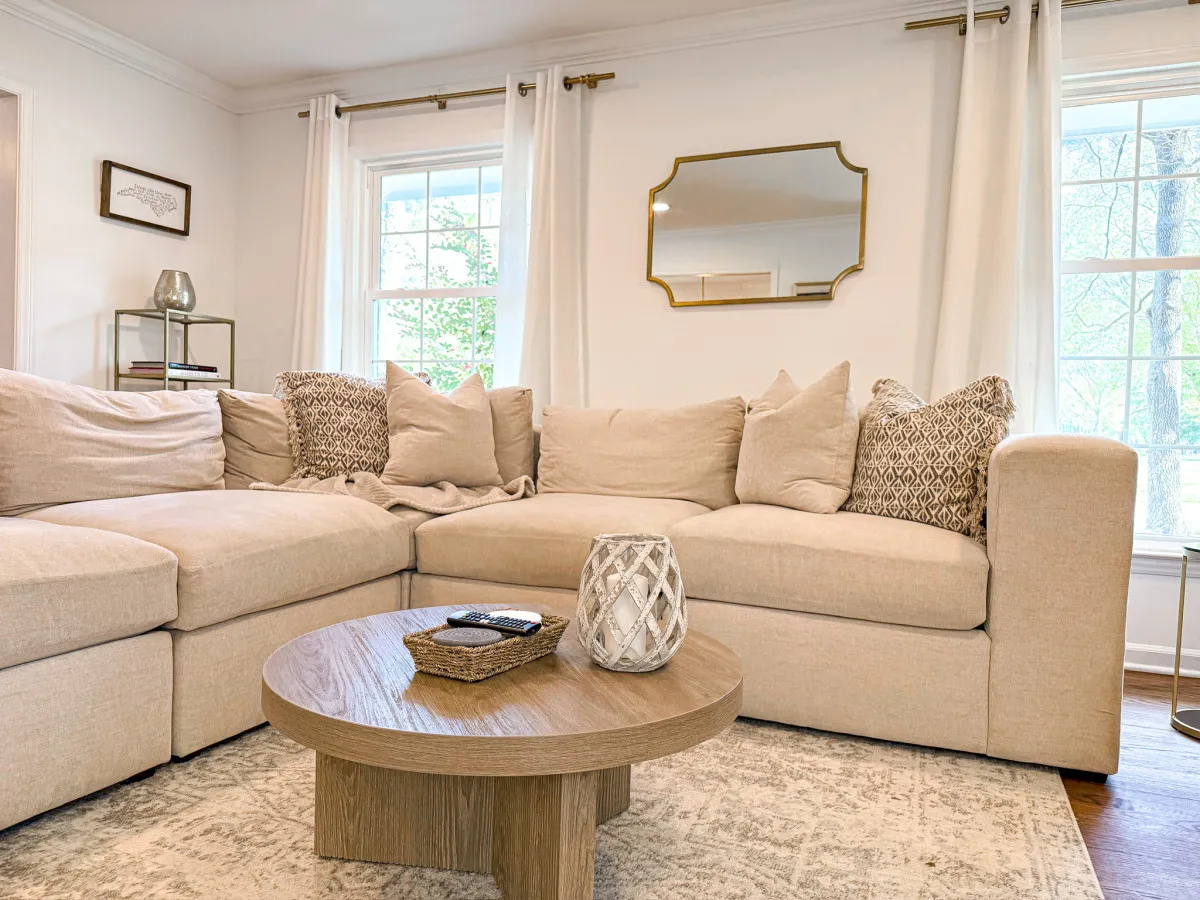 A cozy living area featuring a large sectional sofa, a stylish coffee table, decorative mirror, and windows adorned with curtains. The space is bathed in natural light, creating a welcoming environment ideal for relaxing or socializing during longer stays.